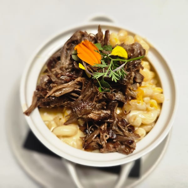 A white bowl of macaroni and cheese topped with shredded beef, garnished with edible flowers and herbs.
