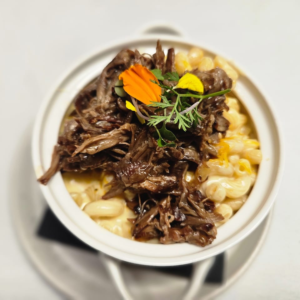 A white bowl of macaroni and cheese topped with shredded beef, garnished with edible flowers and herbs.