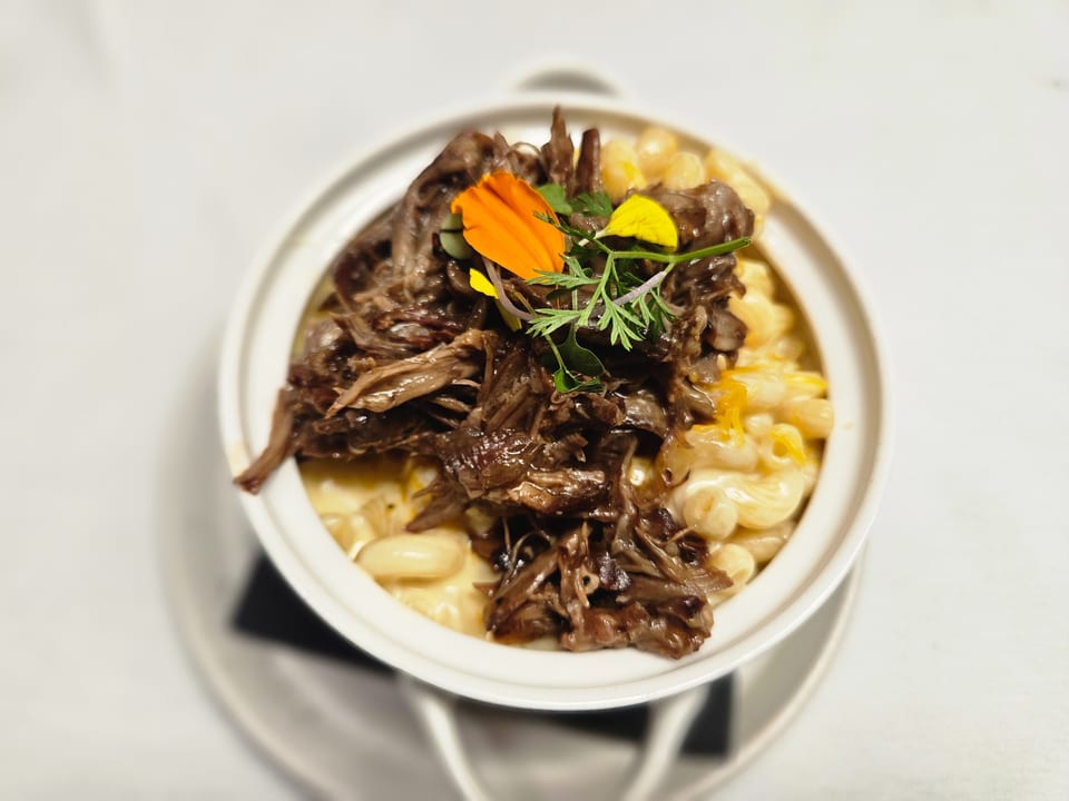 A white bowl of macaroni and cheese topped with shredded beef, garnished with edible flowers and herbs.