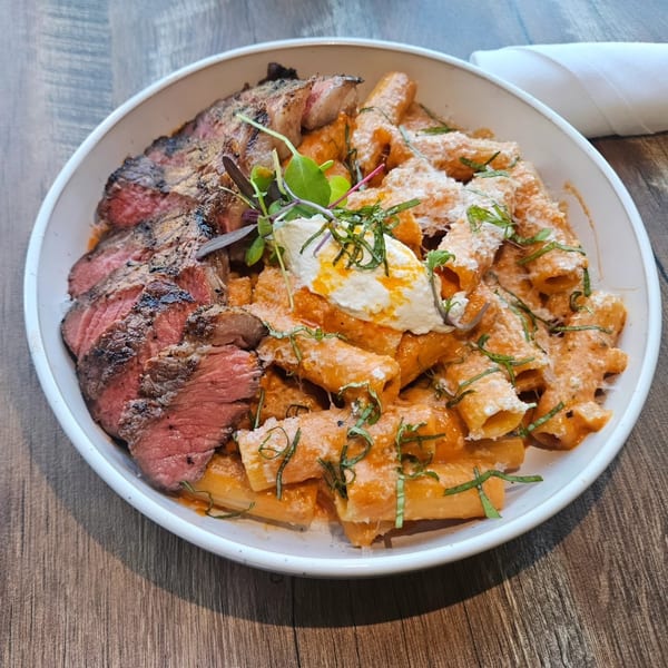 Grilled steak slices and rigatoni pasta with red sauce, cheese, and herbs in a white bowl.