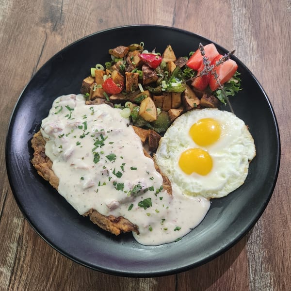 Chicken fried steak with gravy, two fried eggs, roasted potatoes, and fruit on a black plate.