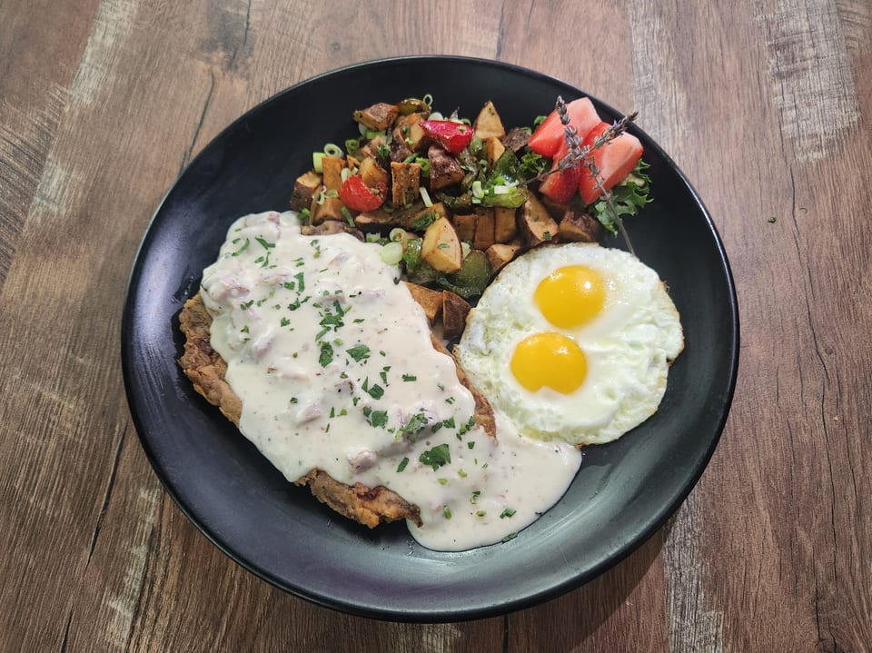 Chicken fried steak with gravy, two fried eggs, roasted potatoes, and fruit on a black plate.