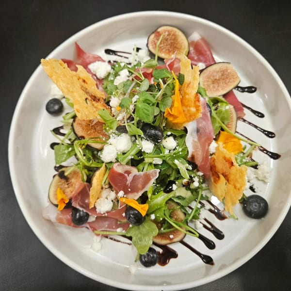 A vibrant salad with prosciutto, figs, blueberries, feta, and greens, drizzled with balsamic glaze.