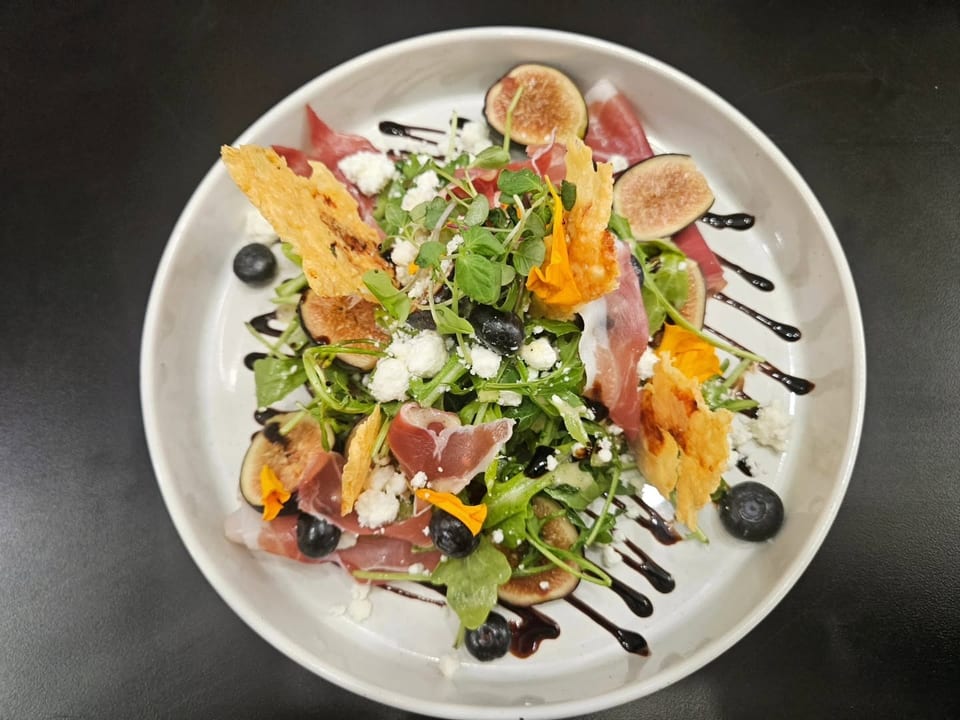 A vibrant salad with prosciutto, figs, blueberries, feta, and greens, drizzled with balsamic glaze.