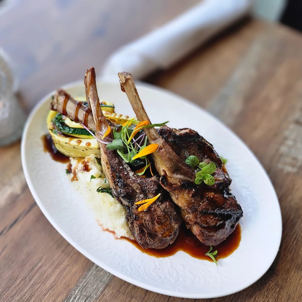 Grilled lamb chops with mashed potatoes, zucchini, and a rich brown sauce on a white plate.