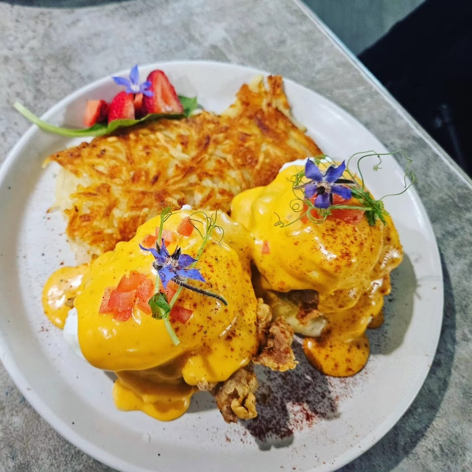 Eggs Benedict with hash browns, garnished with edible flowers and strawberries on a white plate.
