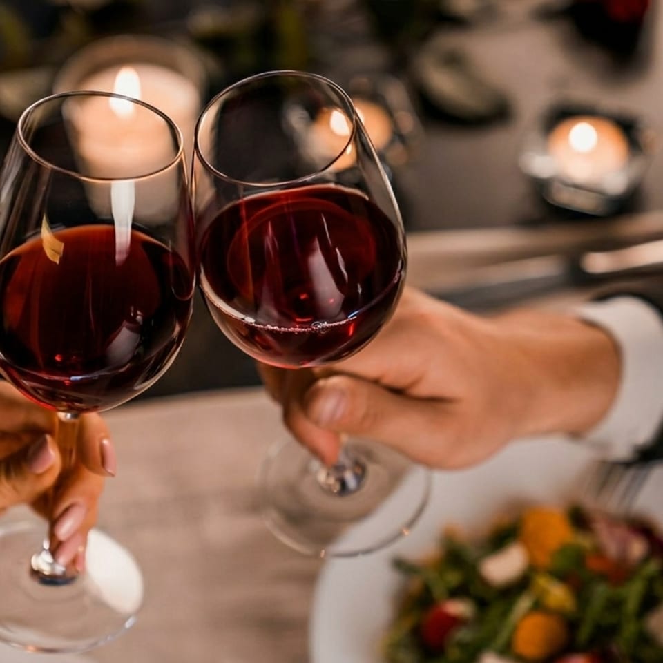 Two people clinking wine glasses over a candlelit dinner with a salad on the table.