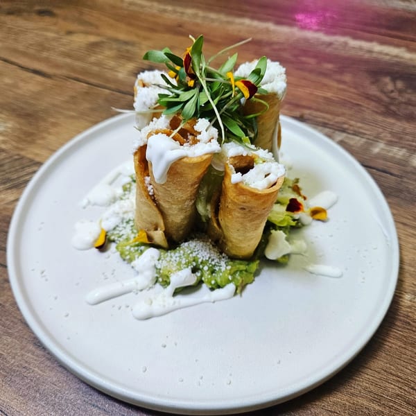 Crispy taquitos with guacamole, sour cream, cotija cheese, and microgreens on a white plate.