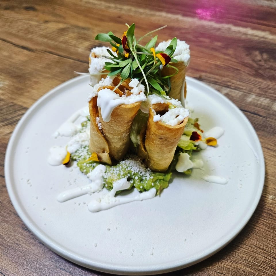 Crispy taquitos with guacamole, sour cream, cotija cheese, and microgreens on a white plate.