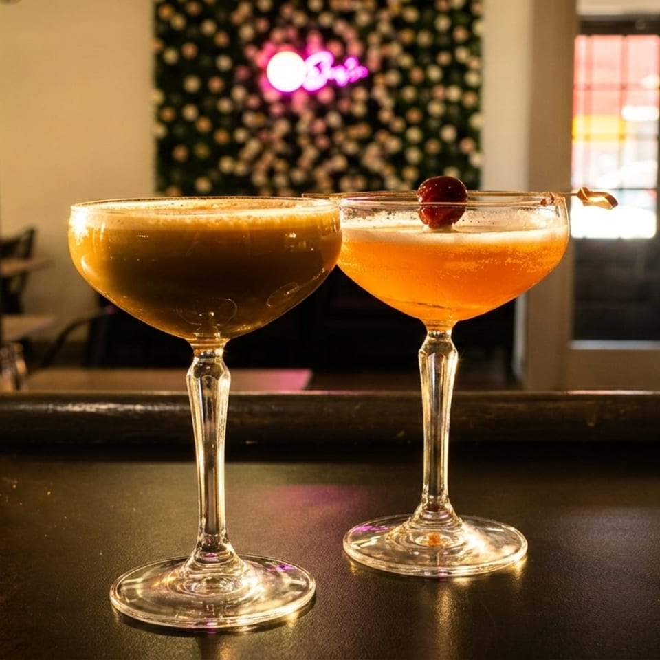 Two cocktails, one dark and one orange with a cherry, on a bar with a neon sign.