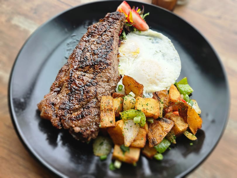 Grilled steak, two fried eggs, and roasted potatoes with peppers and onions on a black plate.