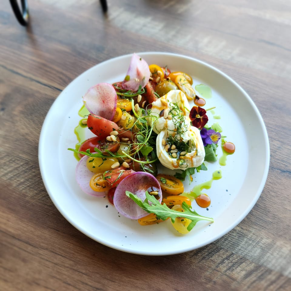 A vibrant burrata salad with colorful tomatoes, radishes, greens, and edible flowers on a white plate.