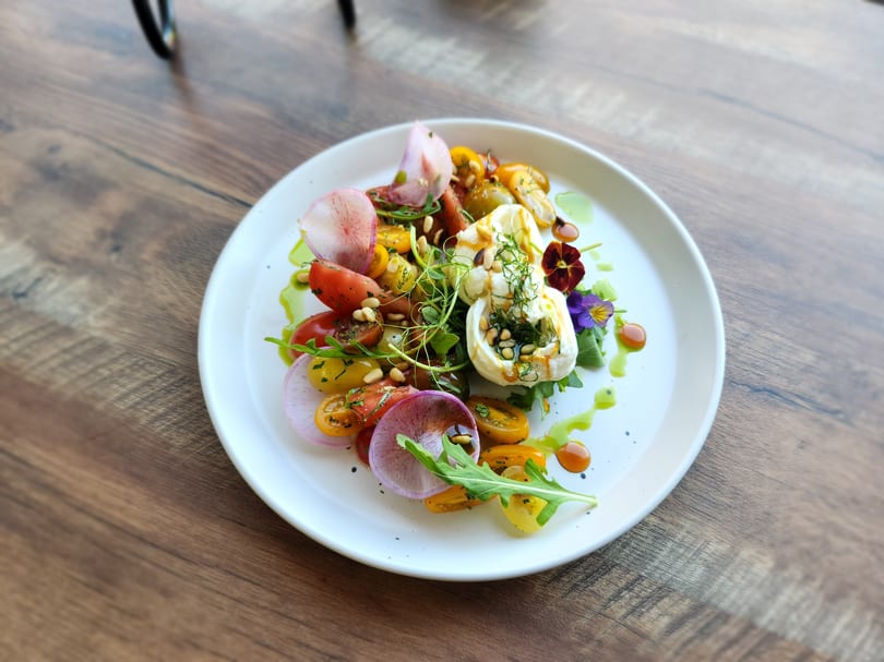 A vibrant burrata salad with colorful tomatoes, radishes, greens, and edible flowers on a white plate.