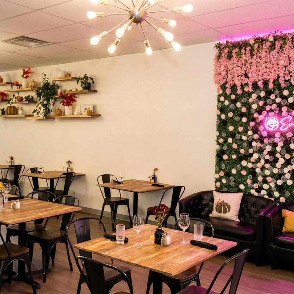 A restaurant interior with wooden tables, black chairs, and a floral wall with a pink neon sign.