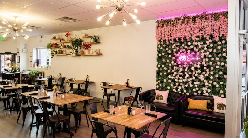 A restaurant interior with wooden tables, black chairs, and a floral wall with a pink neon sign.