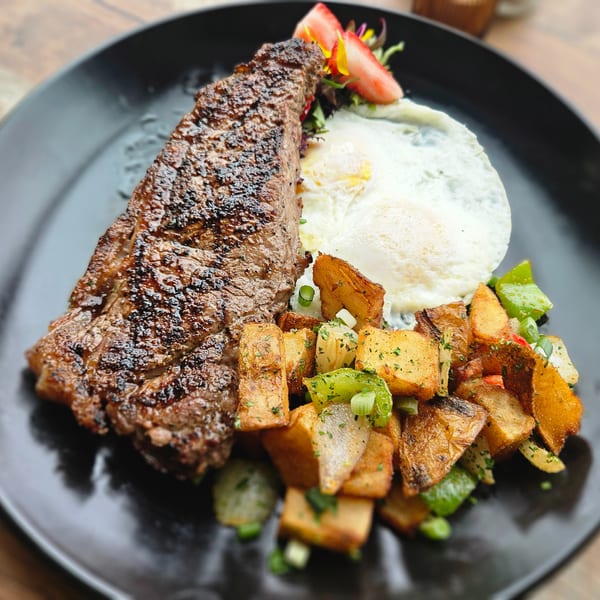 Grilled steak, two fried eggs, and roasted potatoes with peppers and onions on a black plate.